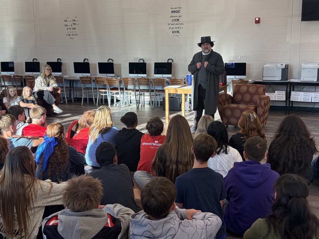 A group of students sitting on the floor listening to a man performing as Edgar Allen Poe.