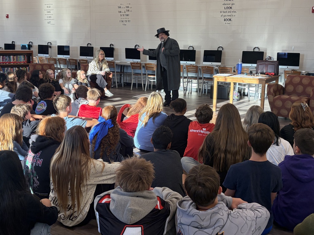 A group of students sitting in the floor watching a performance by a man pretending to be Edgar Allen Poe.