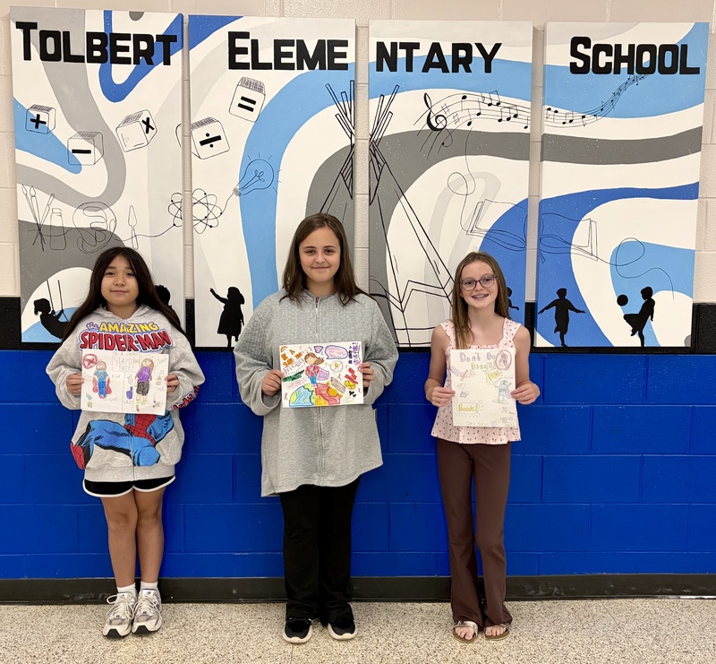 Three students hold their hand-drawn drug prevention posters in front of the blue, white, and gray Tolbert Elementary School mural.