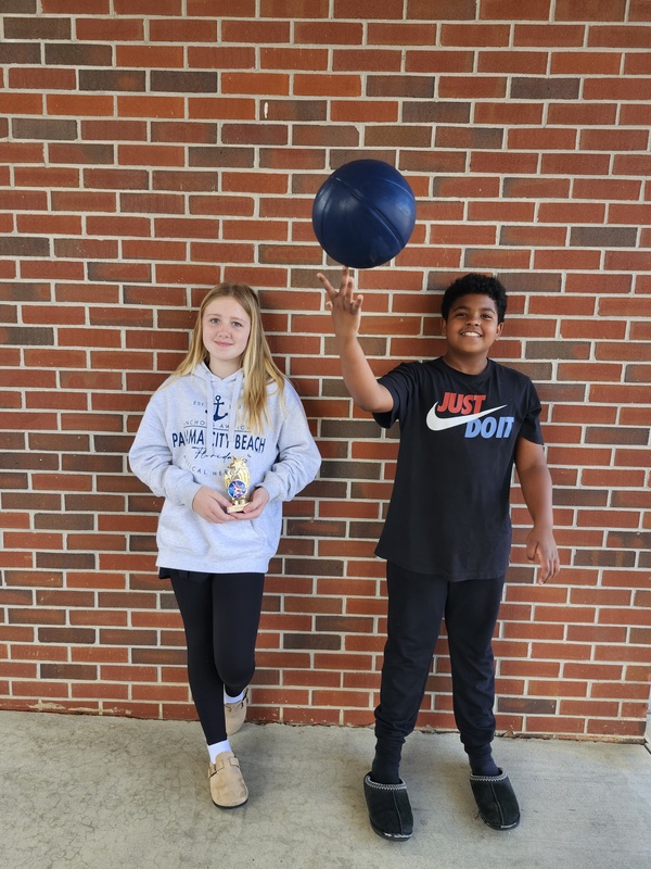 Two students stand against a brick wall; one holds a trophy while the other spins a basketball on their finger.