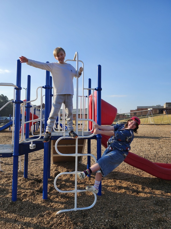 Two students play on a playground structure, one standing on top with arms outstretched while the other hangs from a ladder.