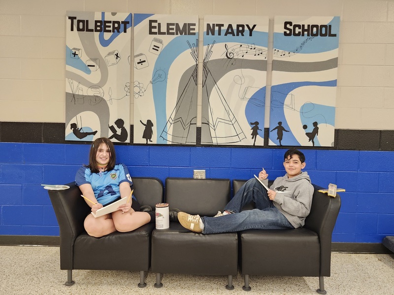 Two students sit on a black couch in front of a “Tolbert Elementary School” mural, smiling and writing on paper.