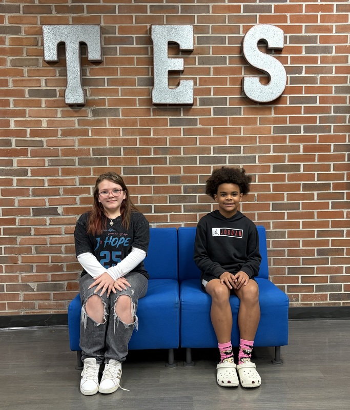 Two students sit on a blue bench in front of a brick wall with large “TES” letters above them.