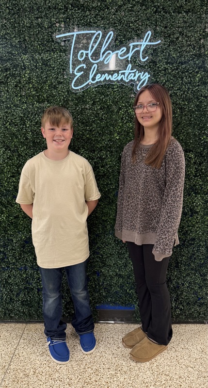 Two students stand in front of a green wall with a “Tolbert Elementary” sign, smiling at the camera.