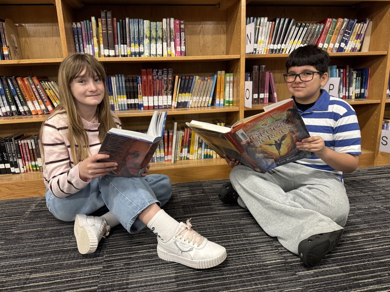 Two students sit on the floor in a library, each holding and reading a book with shelves behind them.