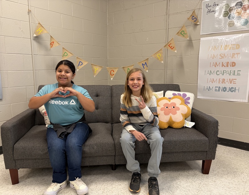 Two students sit on a couch indoors under a “Happy Camper” banner, making hand gestures and smiling.