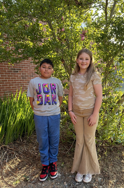 Two students stand side by side outdoors near plants and a tree, smiling at the camera.