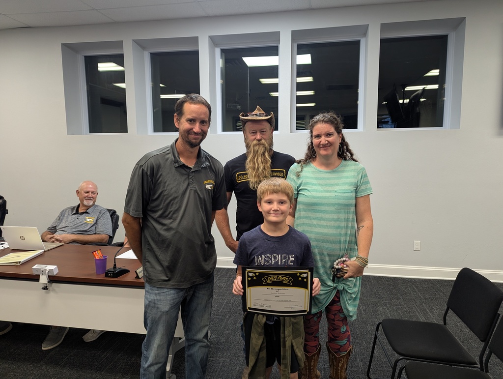 A young boy stands smiling and holding a certificate in front of three adults during a recognition event in a meeting room. The boy’s parents stand beside him, and two men from the school district stand nearby, one wearing a Grandview Eagles shirt. In the background, another man sits at a table with a laptop, smiling.