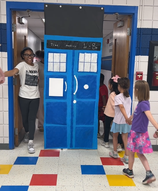 An image of first graders walking behind a door holding hands and eigth grade students walking out the other side pretending to be in a time machine