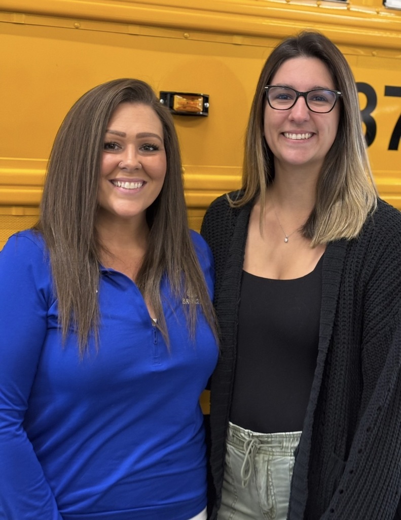 On the left, Kristine Coley-Thomson and on right, Nicole Bishop, both standing in front of the side of a school bus