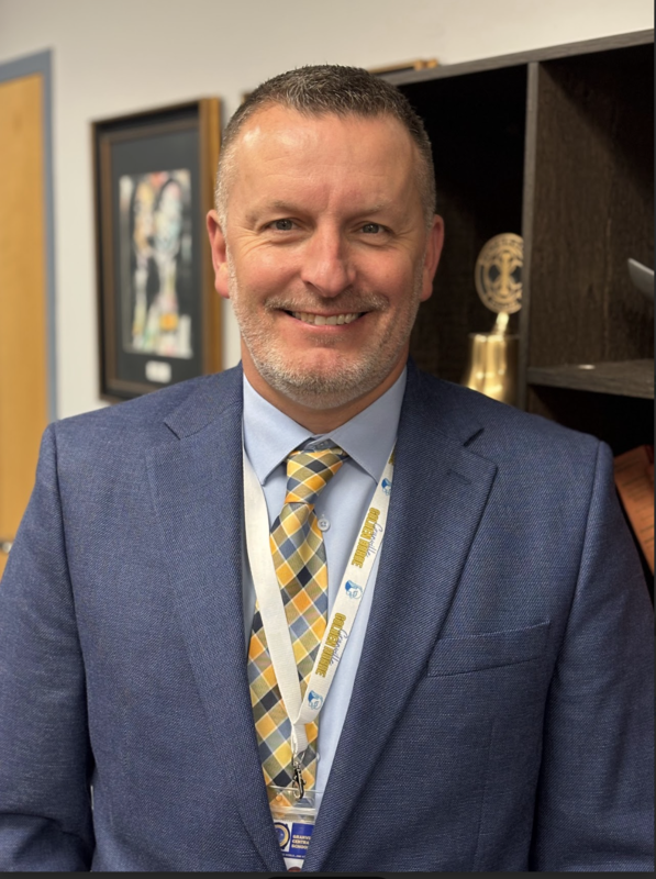 Mr. Hall, wearing a blue suit jacket and a blue and gold tie, standing in the District office conference room. 