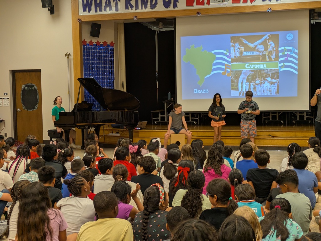 Three students play percussion instruments, a pianist plays a grand piano, and many students watch in the audience. 