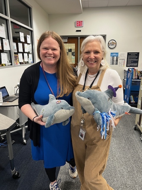 January SAM recipients Mrs. Hill wearing a blue dress and a black sweater. Mrs. Gray wearing tan overalls and a white shirt. Both are holding stuffed dolphins and they are standing in the library smiling.