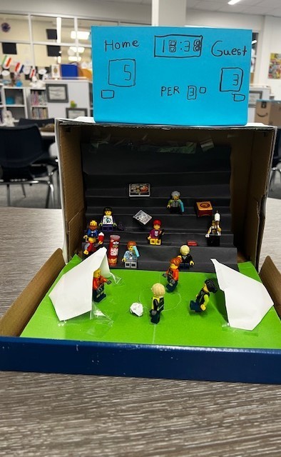 cardboard box with miniature lego people. some are on a paper soccer field and some are sitting in the stands