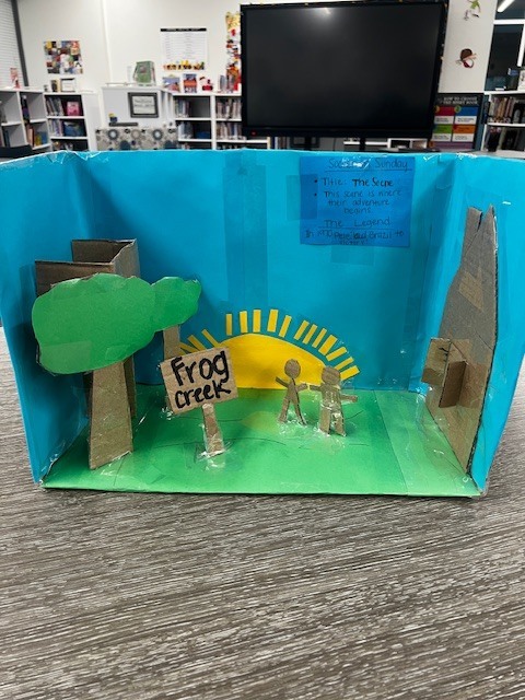 cardboard box with blue background and green bottom. cardboard tree, house, two children, and a sign that says "frog creek"