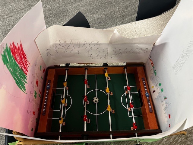 miniature foosball table with paper around it. On the paper there are people drawn as if they are fans in the stands.