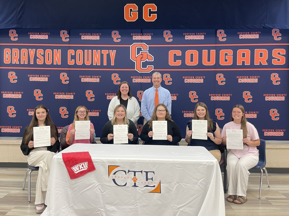 Students and teachers hold certificates in front of a Grayson County Cougars step and repeat banner at an education signing day event