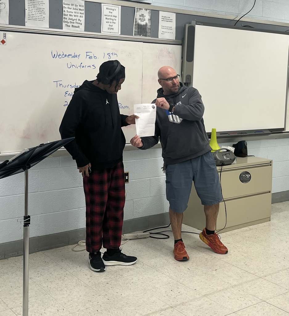 Two men stand in a classroom; one holds a paper, and the other holds a microphone. A whiteboard with writing is behind them.