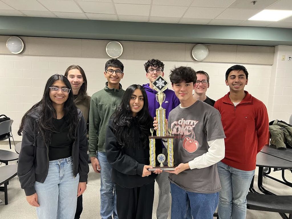 A group of students holding a trophy in a classroom with tables and chairs. One student holds the trophy.