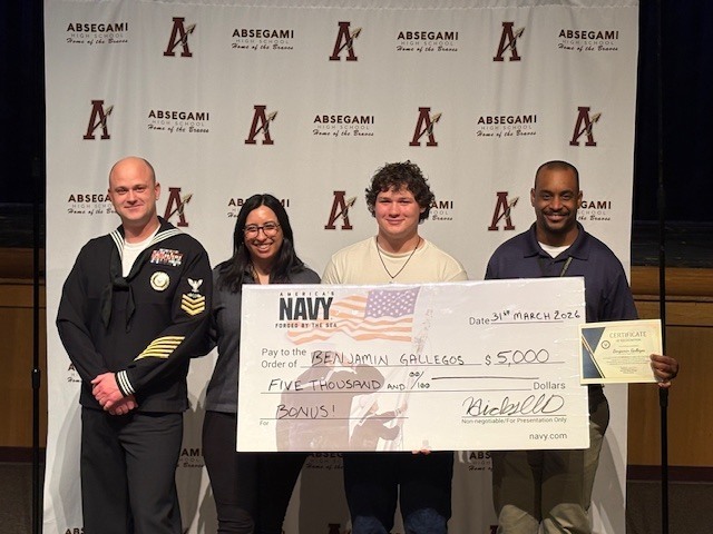 Four adults stand together holding a large check and awards. A large white banner with the letter A in various colors is behind them.