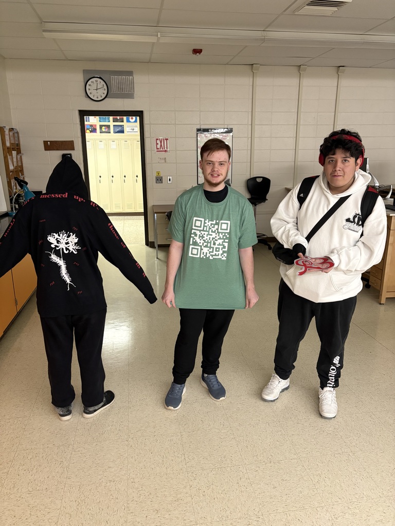 Three people stand in a classroom. One wears a black hoodie, another a green T-shirt, and the third a white hoodie.