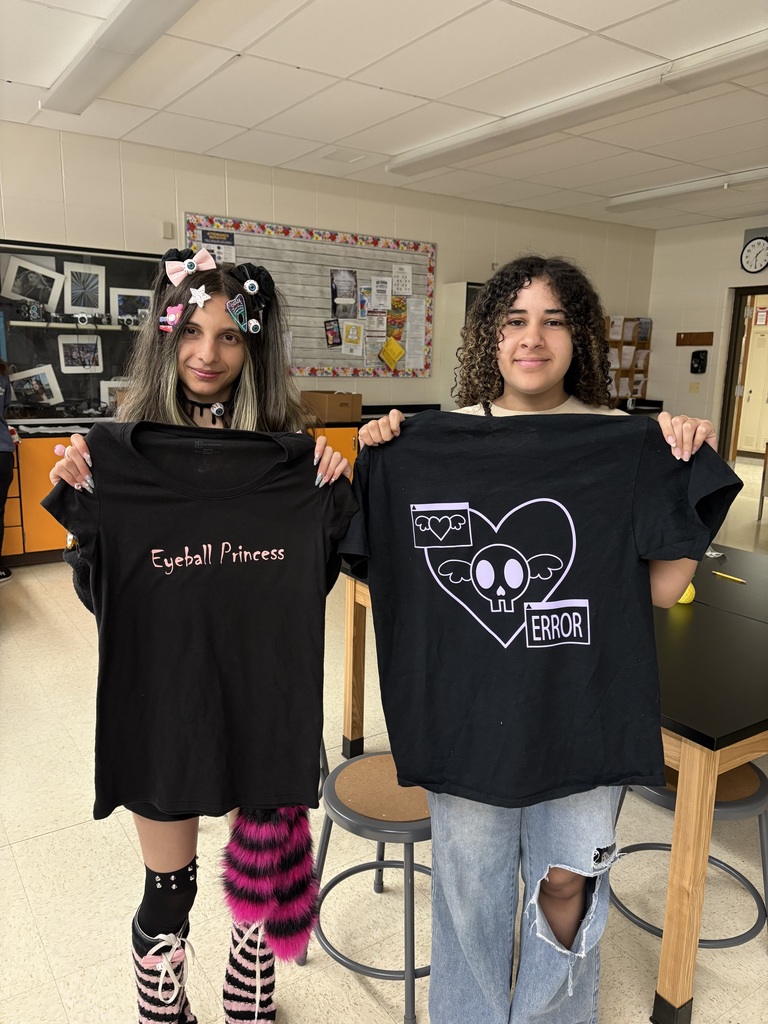 Two individuals in a classroom with white walls, one holds a shirt reading "Eyeball Princess," and the other holds a shirt with a heart design and "ERROR" text.