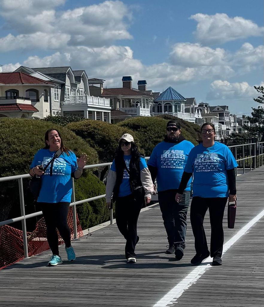 Four people in blue t-shirts walk on a boardwalk, with houses and greenery behind them.