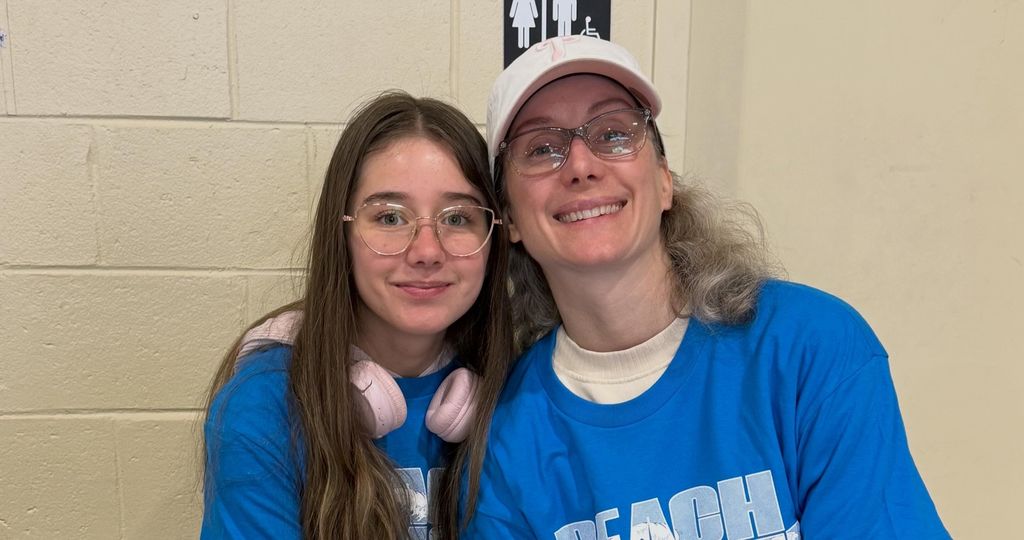 Two women in blue t-shirts, one wearing a white cap, standing close to each other and smiling.