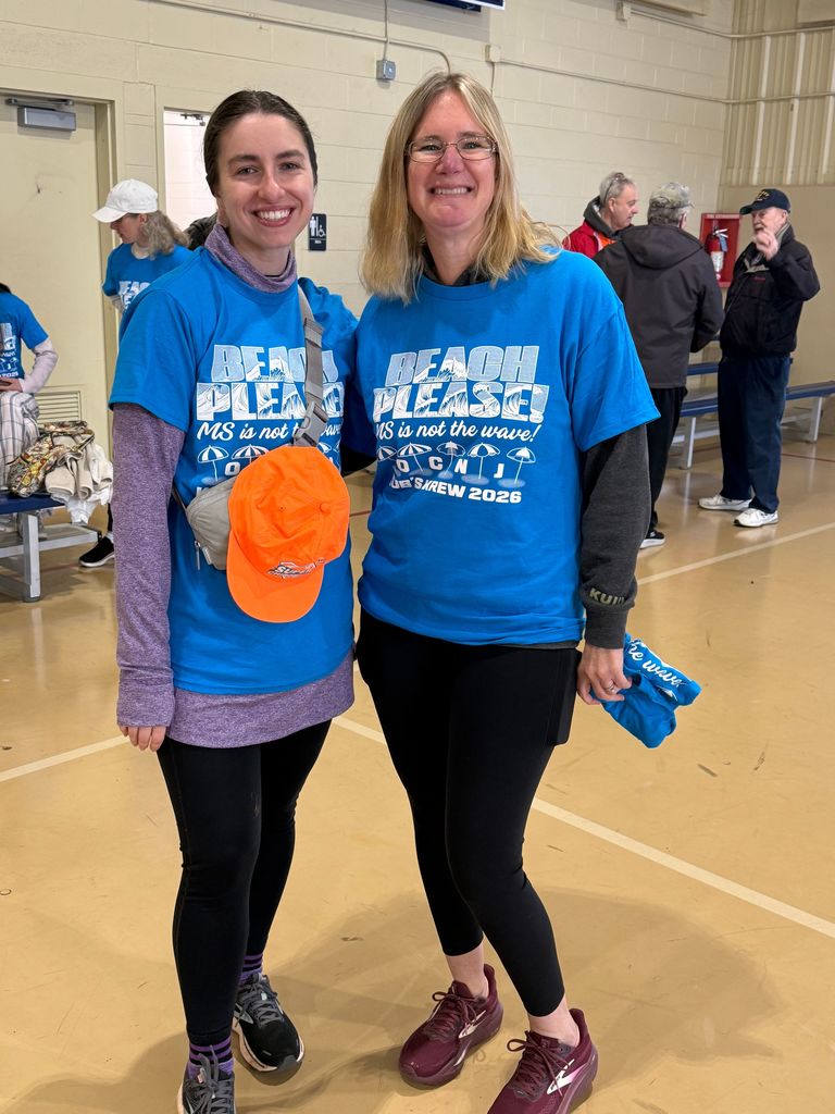 Two women in matching blue shirts, one carrying a hat, smile and pose in a room.