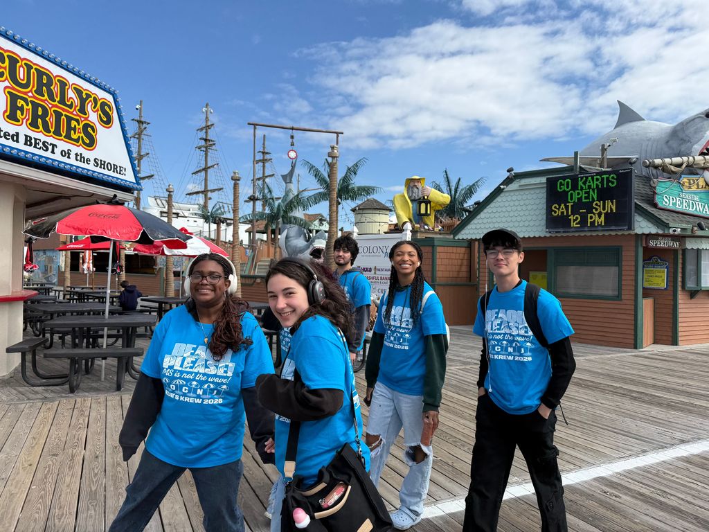 Five people in blue shirts and sneakers stand on a boardwalk in front of a restaurant.