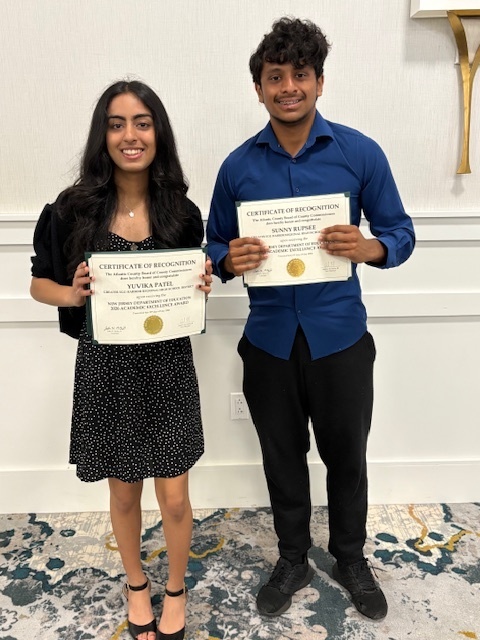 A woman and a man hold certificates in front of a white wall with a patterned rug.