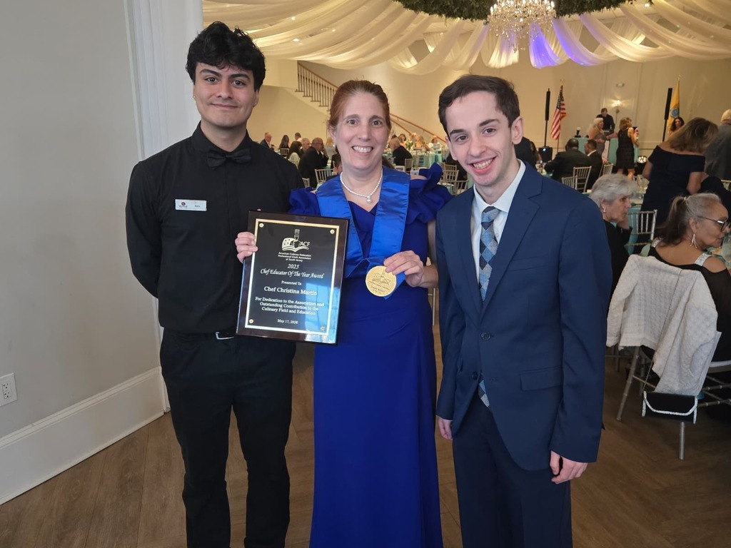 Three people pose with a plaque and medal in a room with a wooden floor, white walls, and a ceiling with lights.