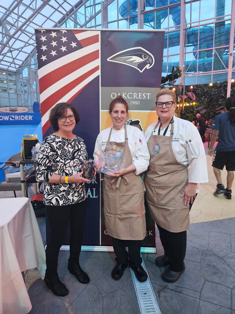 Three women pose next to a banner featuring an American flag and an eagle. One holds an award.