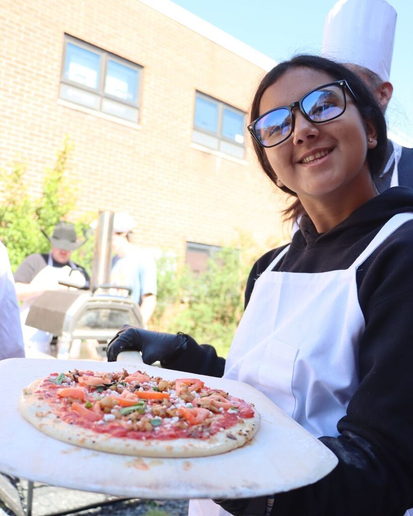 Person in a white apron holding a pizza with vegetables and red sauce on a cutting board.