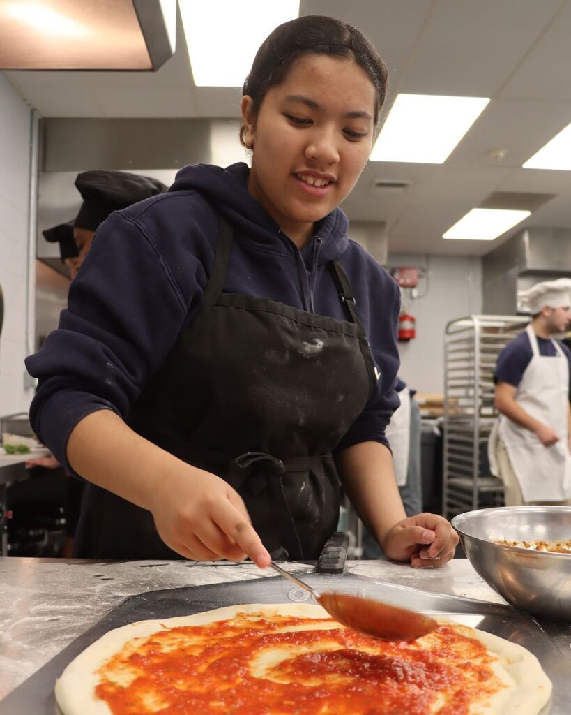 Woman wearing an apron, using a spoon to spread sauce on a pizza base.