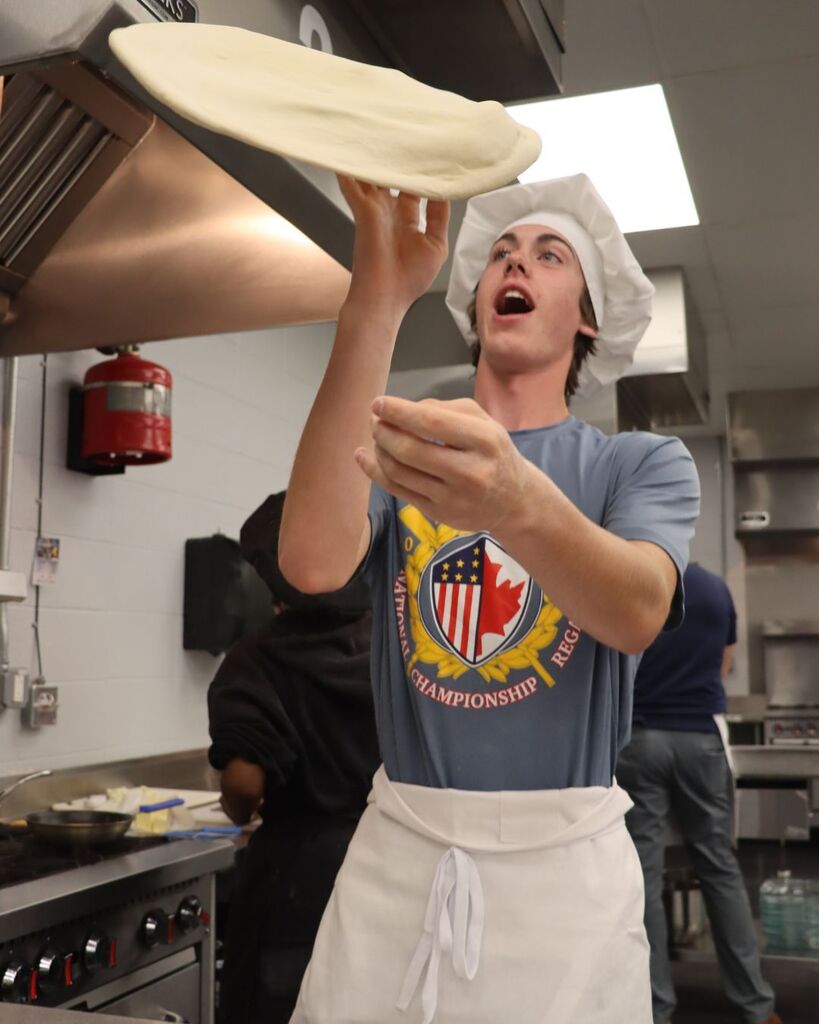 A chef tosses a pizza in the air in a kitchen. He wears a chef's hat and apron.