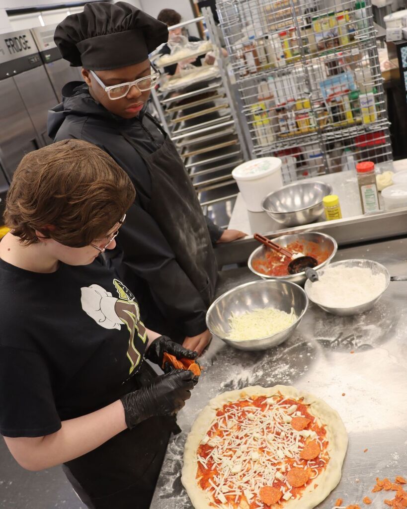 Two individuals with glasses prepare a pizza topped with cheese and pepperoni in a commercial kitchen.