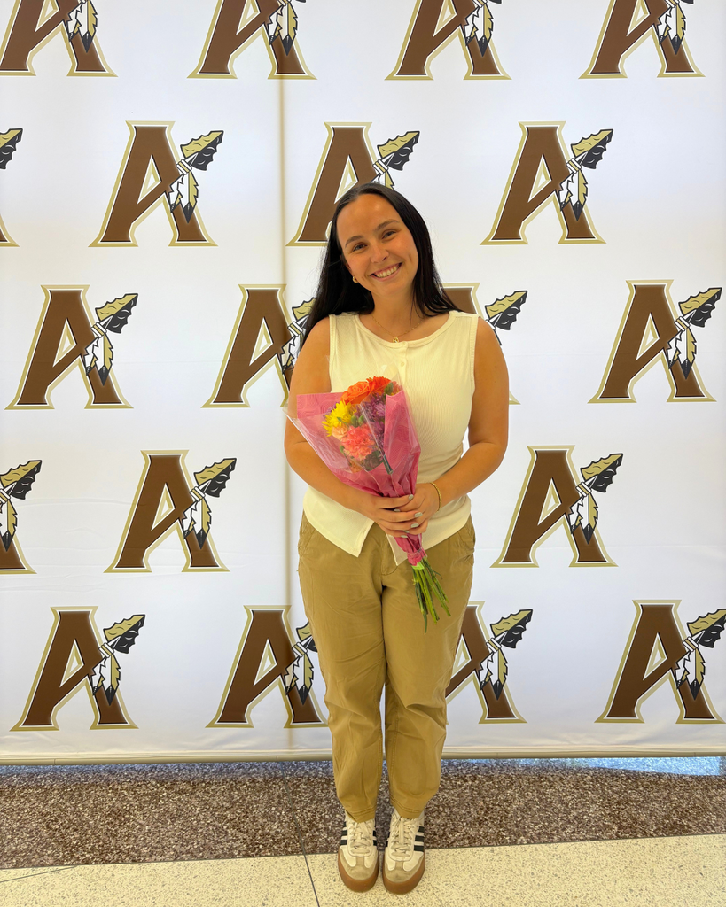A woman in a white sleeveless top and khaki pants stands in front of a wall with the letter A. She holds a bouquet of flowers.