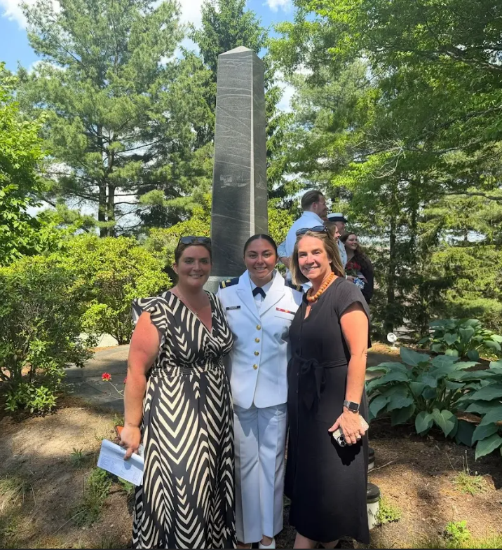 Three people stand next to a stone monument, one in a military uniform, among trees and plants.