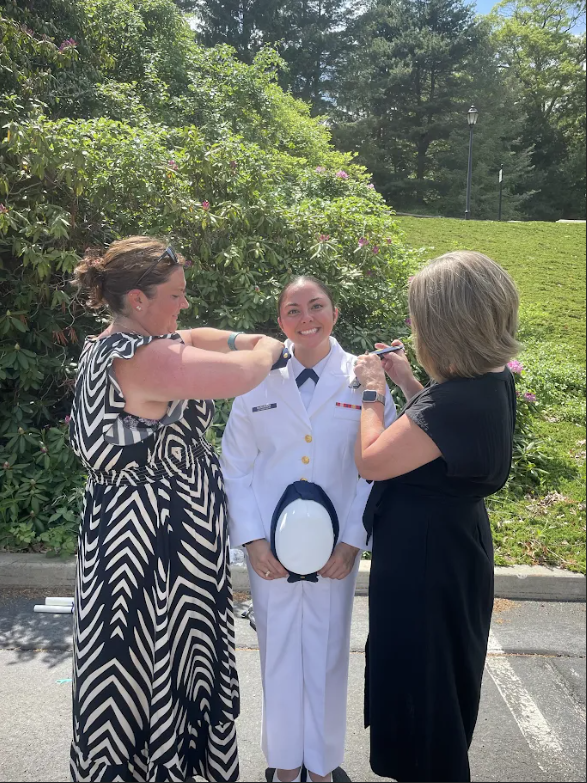 A woman in a coast guard uniform has her cap pinned on by two others. They stand in a parking lot with greenery.