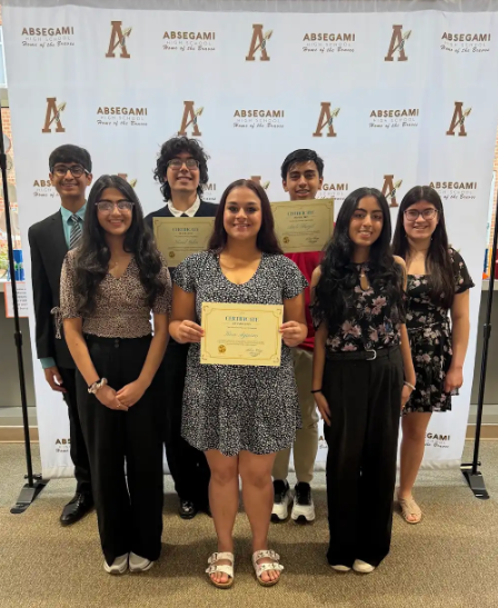 Six students in casual attire stand in front of a white banner with the letters A and ABSEGAMI. A girl holds a yellow certificate.