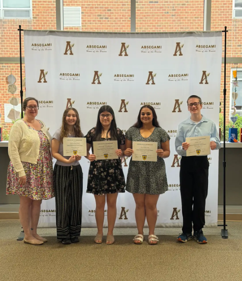 Five people stand together holding certificates. A large white backdrop with the letters A and ABE behind them.