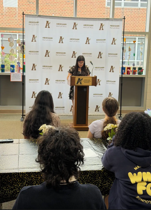 A woman speaks at a podium with a banner behind her, addressing a group of people seated at tables.