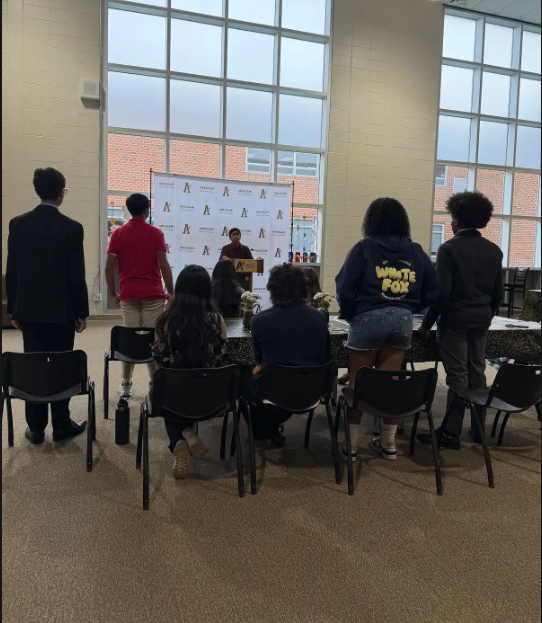 People gather around tables inside a room with large windows. Some individuals stand near a banner.