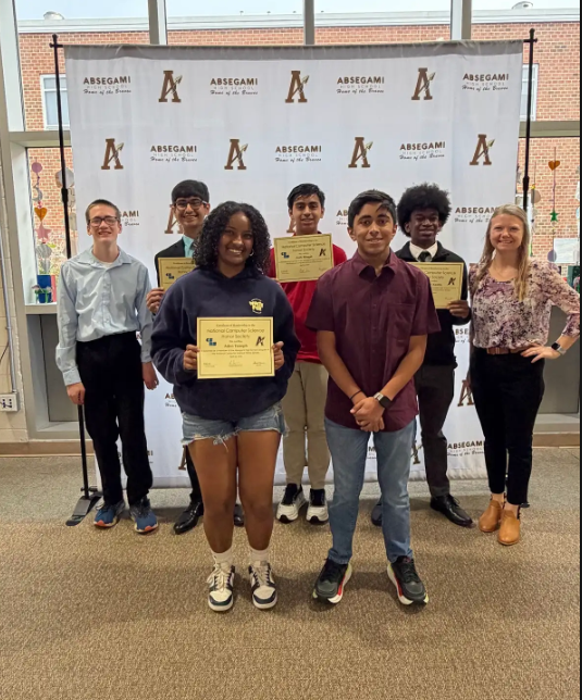 Group of students holding certificates. They stand in a row, smiling, with a banner behind them.