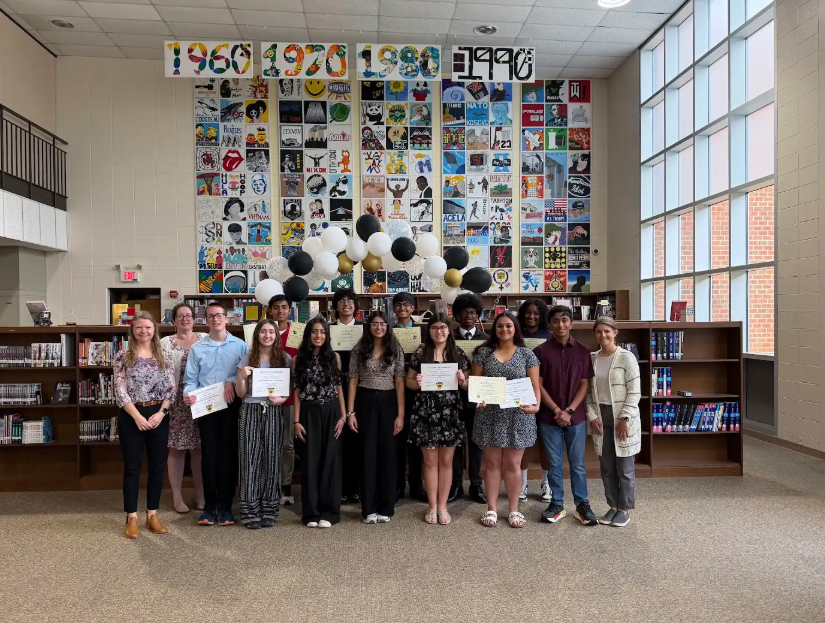 A group of individuals holding certificates stands in a library with bookshelves and a colorful wall.