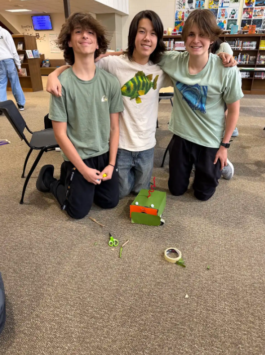 Three boys, two with arms around each other, kneeling next to a small green box, with a chair and books in the background.