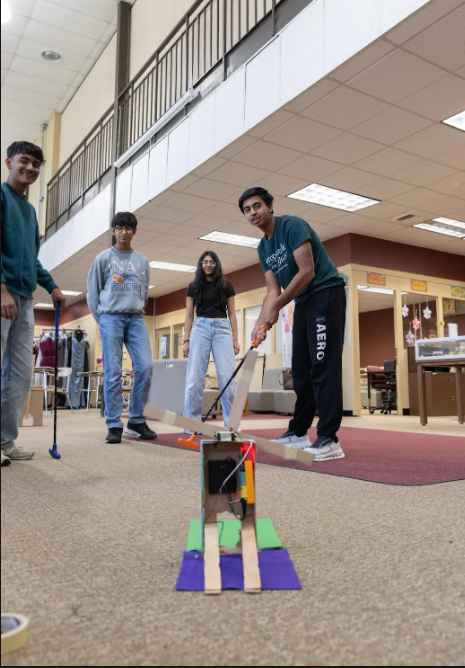Four people inside a hallway, one playing golf with a robot, others watching. The hallway has a carpet, a desk, and a railing.