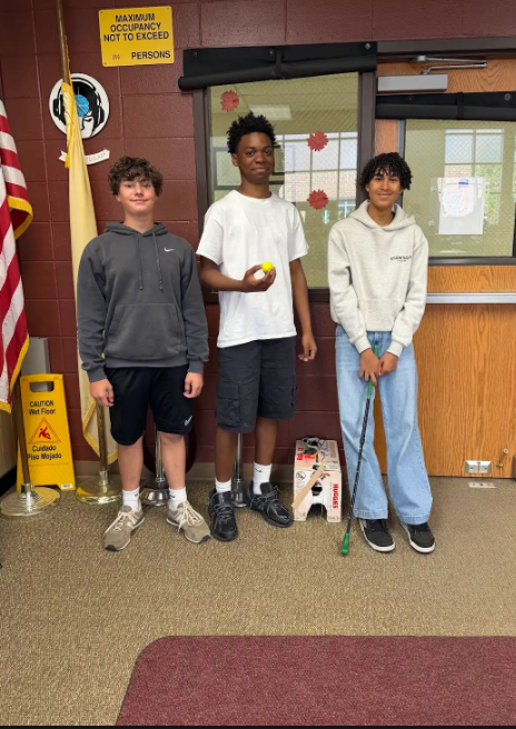Three boys stand in a room with a flag, one holding a golf club and ball.