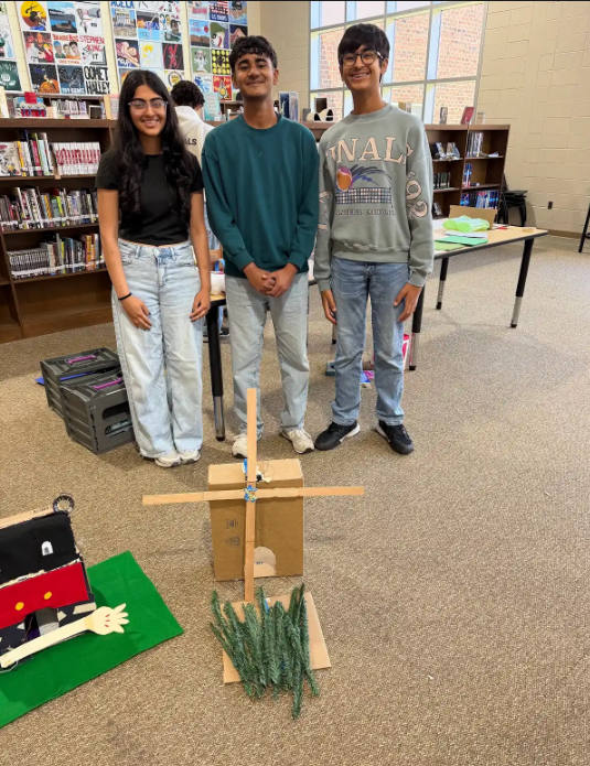 Three students stand in a classroom with a homemade windmill and a small, decorated object on a table.